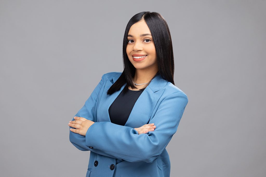 Headshot of REMAX of Reading real estate agent Alexa Henriquez wearing a blue jacket on a grey background. 