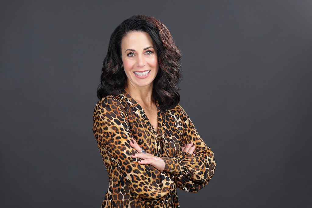 Headshot of a smiling woman with dark hair wearing a leopard print shirt on a dark neutral background. 