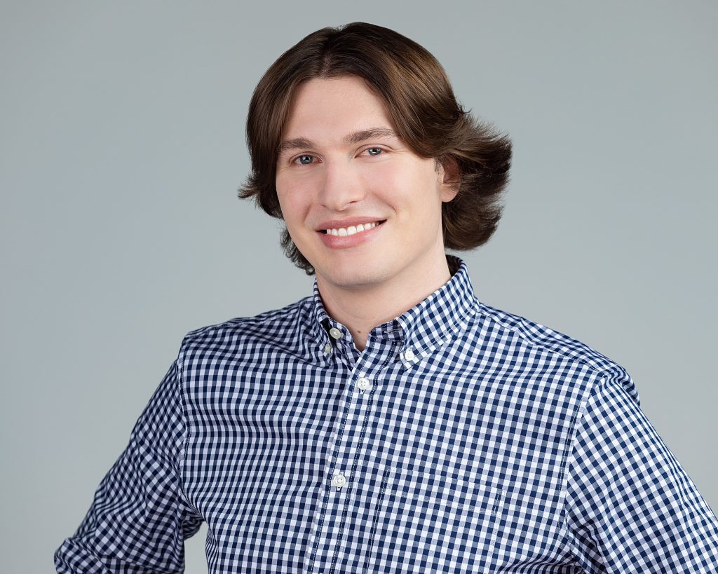 Young man wearing plaid shirt on neutral background smiles for his staff headshot. 