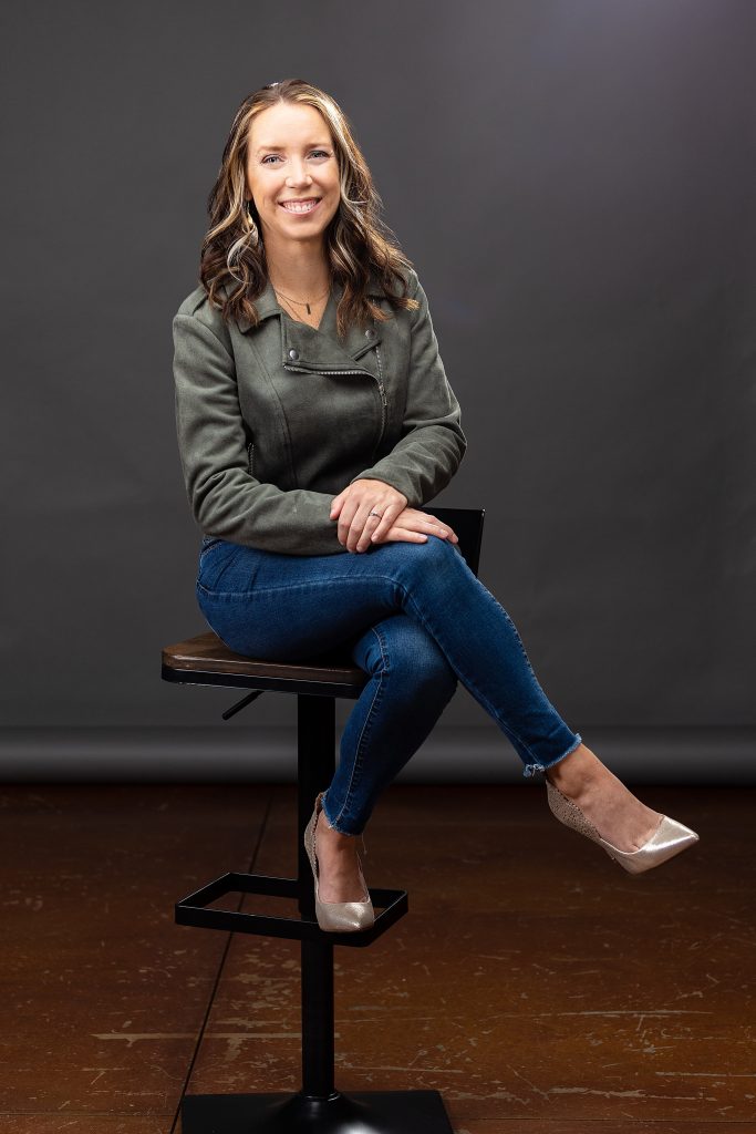 Professional business headshot of a smiling woman sitting on a tall chair with legs crossed in Berks County photo studio. 