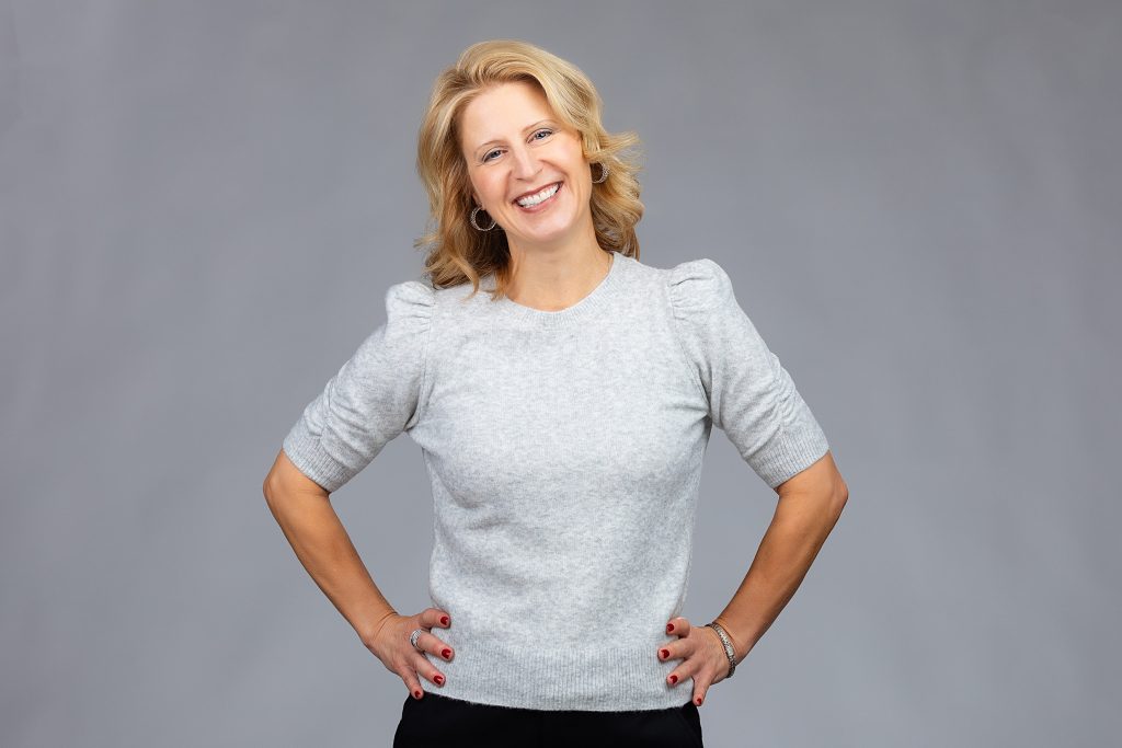 Social profile pic update to a professional headshot: business woman with blond hair on a light grey background, smiling, with hands on hips. 