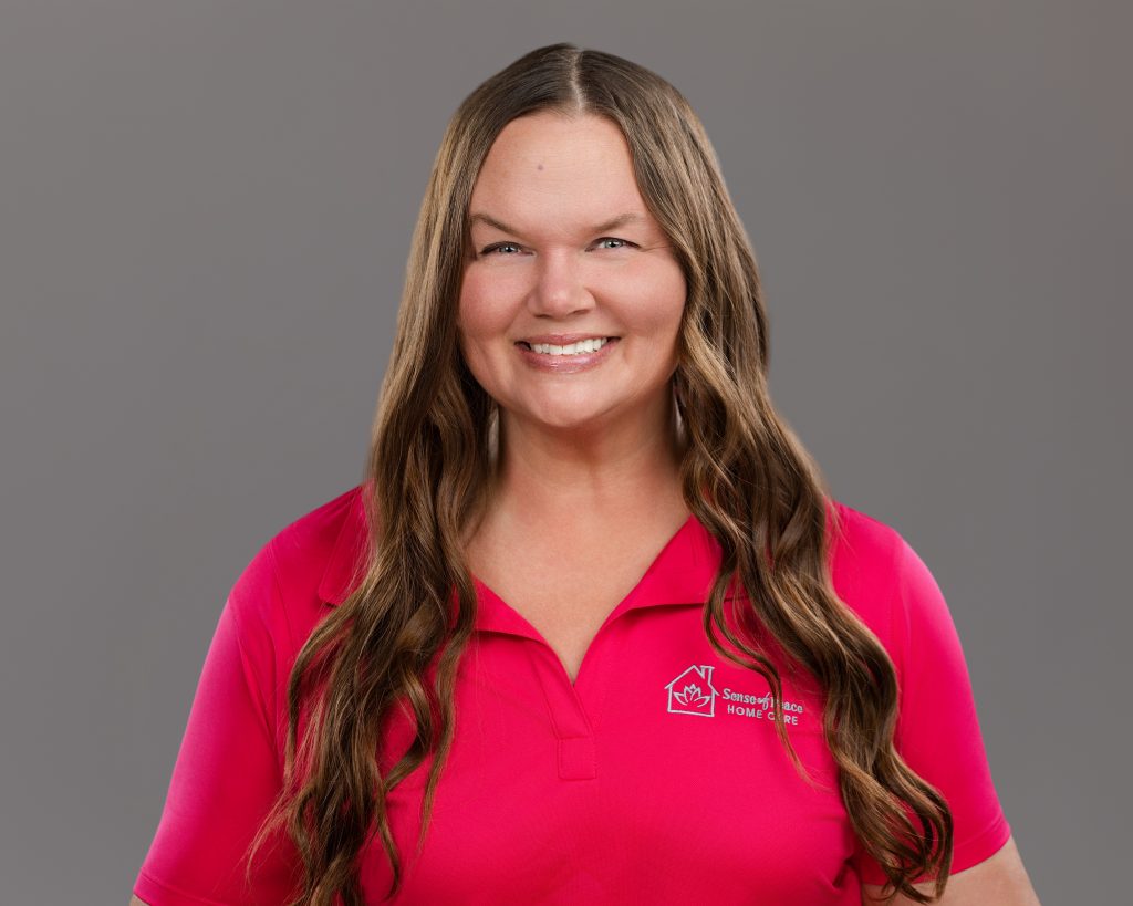 Home care staff photo of caregiver with long brown hair and a pink polo shirt on grey background