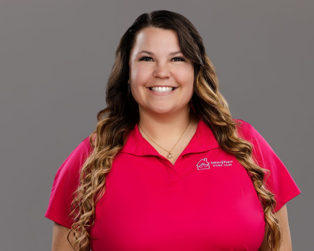 Home care staff photo of caregiver with long brown hair and a pink polo shirt on grey background