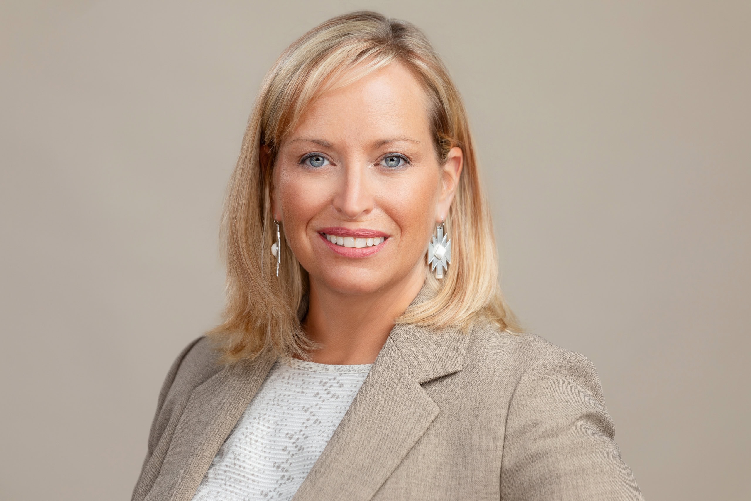 Headshot of a smiling business woman with sparkly earrings and tan jacket, on a warm background.