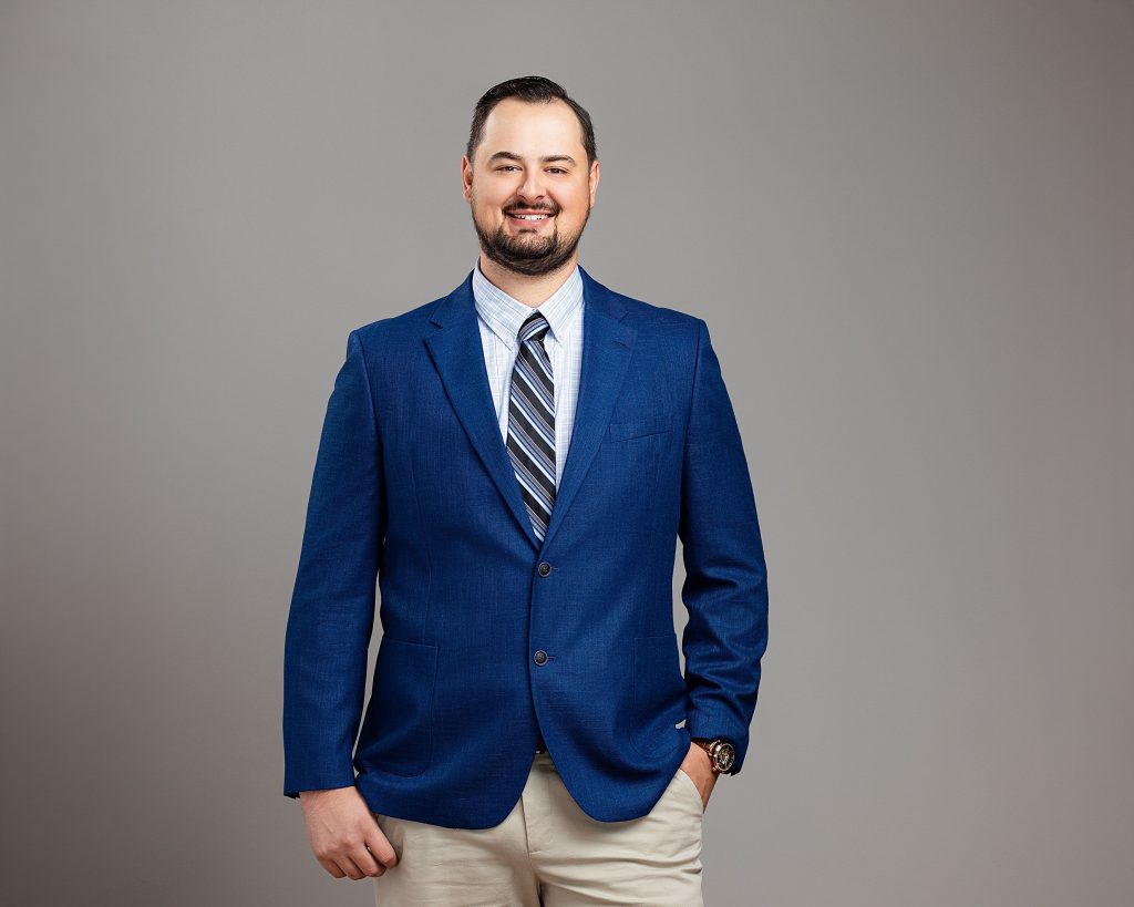 Professional portrait of a smiling man in a blue suit jacket against a modern grey background