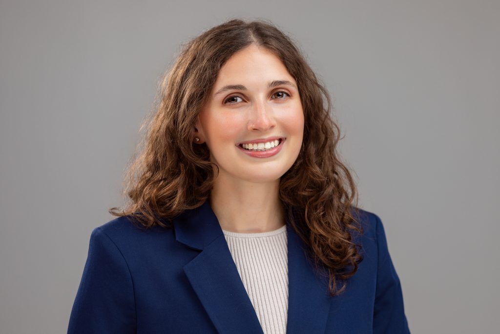 Headshot of a medical student who is applying for residency. The woman has long brown hair and is smiling gently. There is a neutral grey background. 