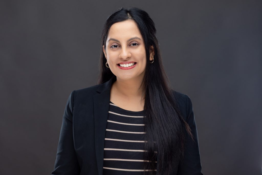 Headshot of a smiling doctoral candidate with dark hair and dark shirt against a dark backdrop. 