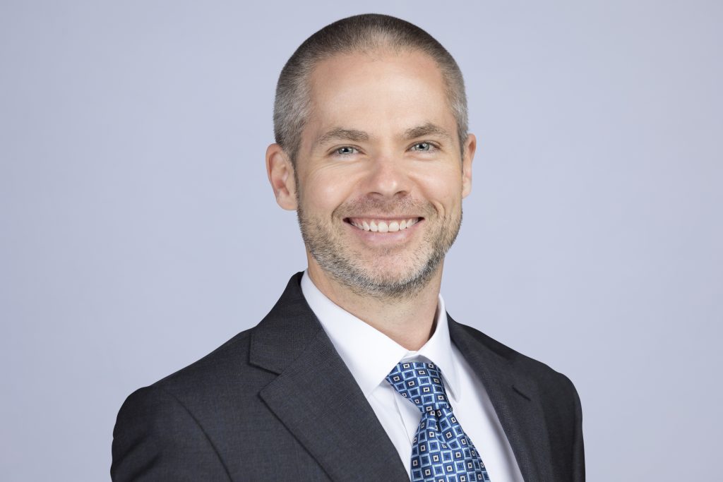 Headshot of Ben Tercha, cybersecurity expert, smiling and wearing a black jacket and blue tie against a light blue background. 
