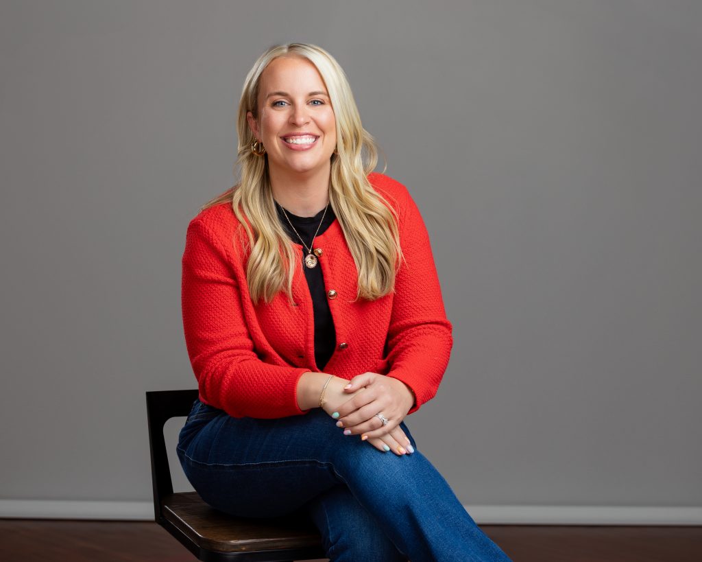 Headshot of a real estate agent wearing a red sweater,seated on a chair, smiling and engaged with the viewer. 