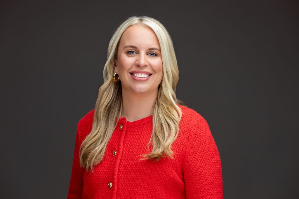 Headshot of a smiling blond real estate agent wearing a red sweater against a dark background. 