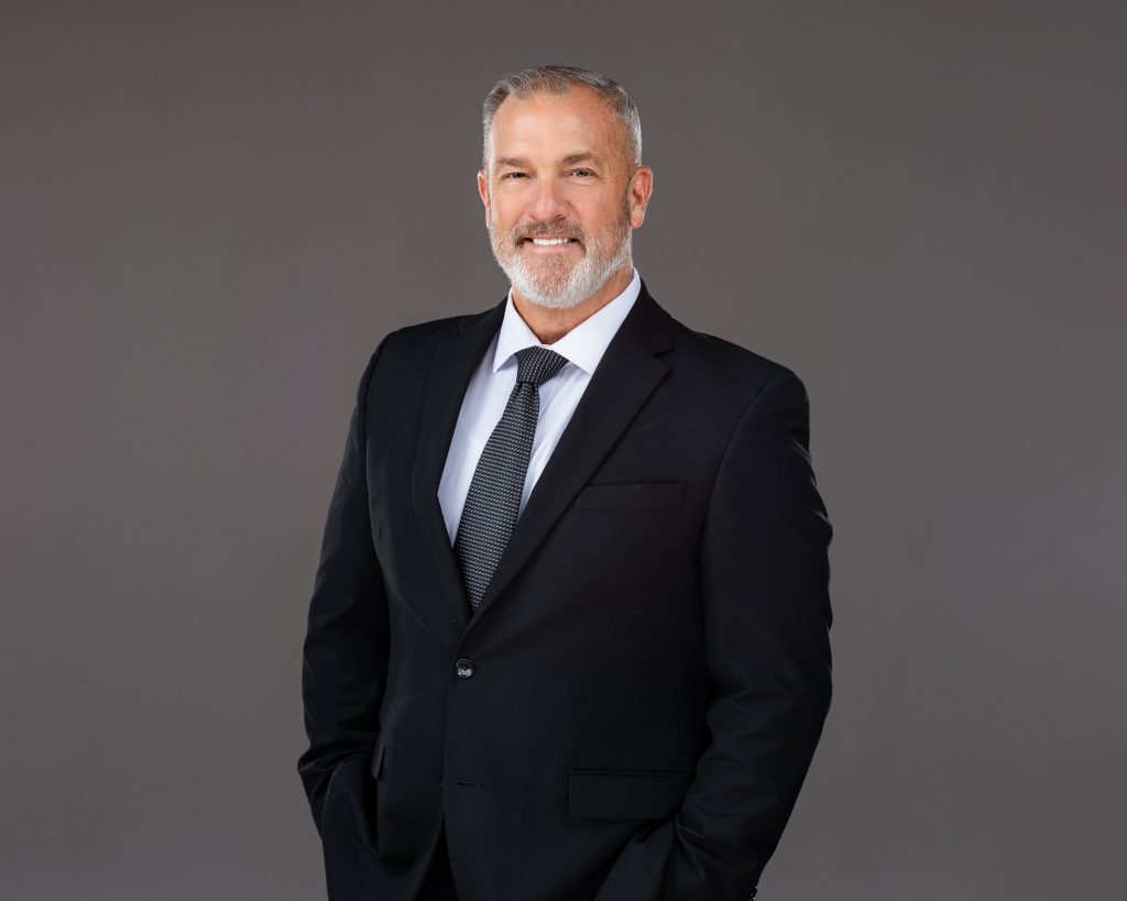 A waist-up headshot of a businessman in a black suit jacket and black tie on a dark grey background. 