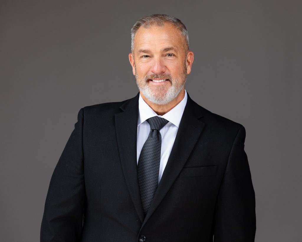 Business headshot of a businessman in a black suit jacket and black tie, smiling. 