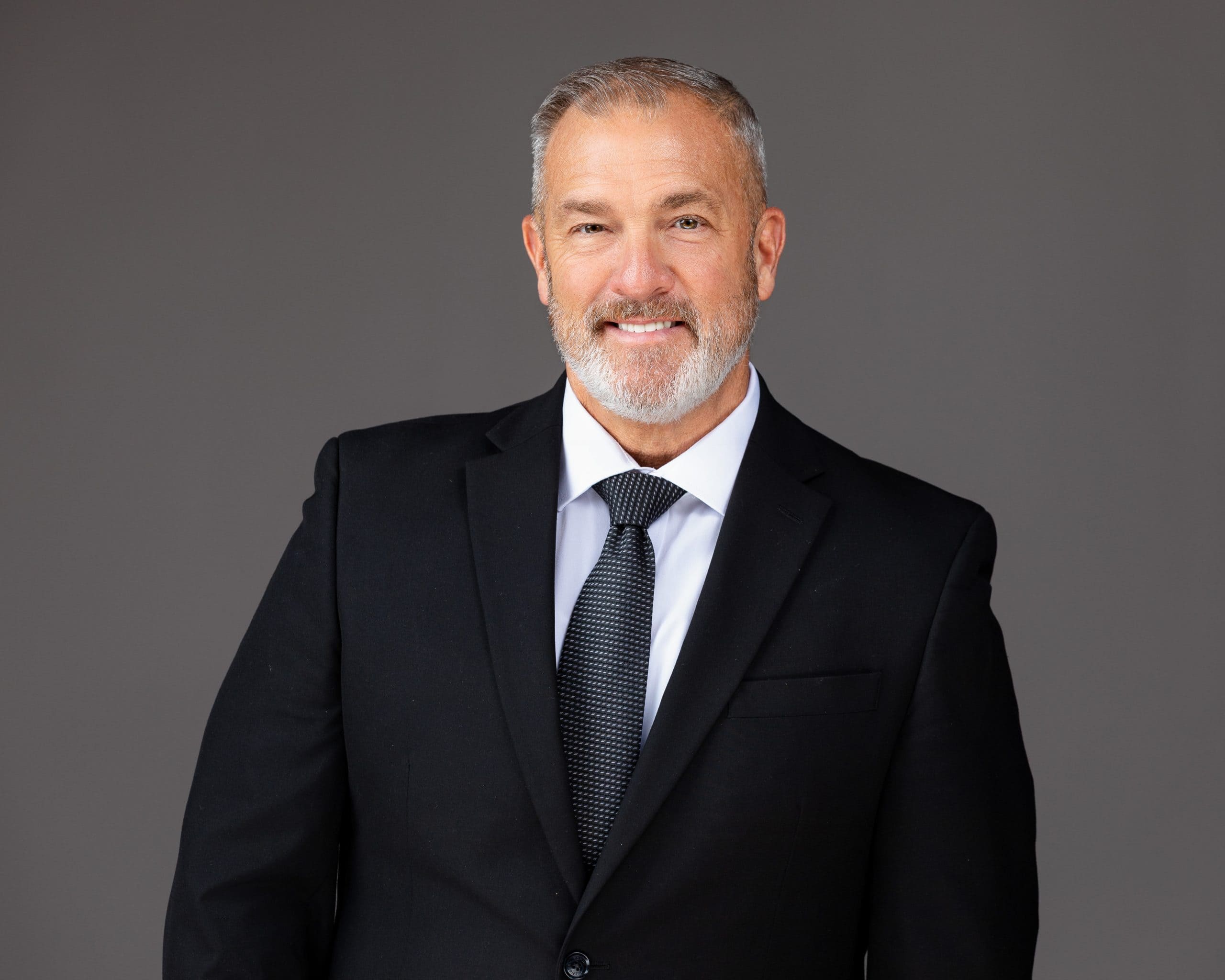 Business headshot of a businessman in a black suit jacket and black tie, smiling.