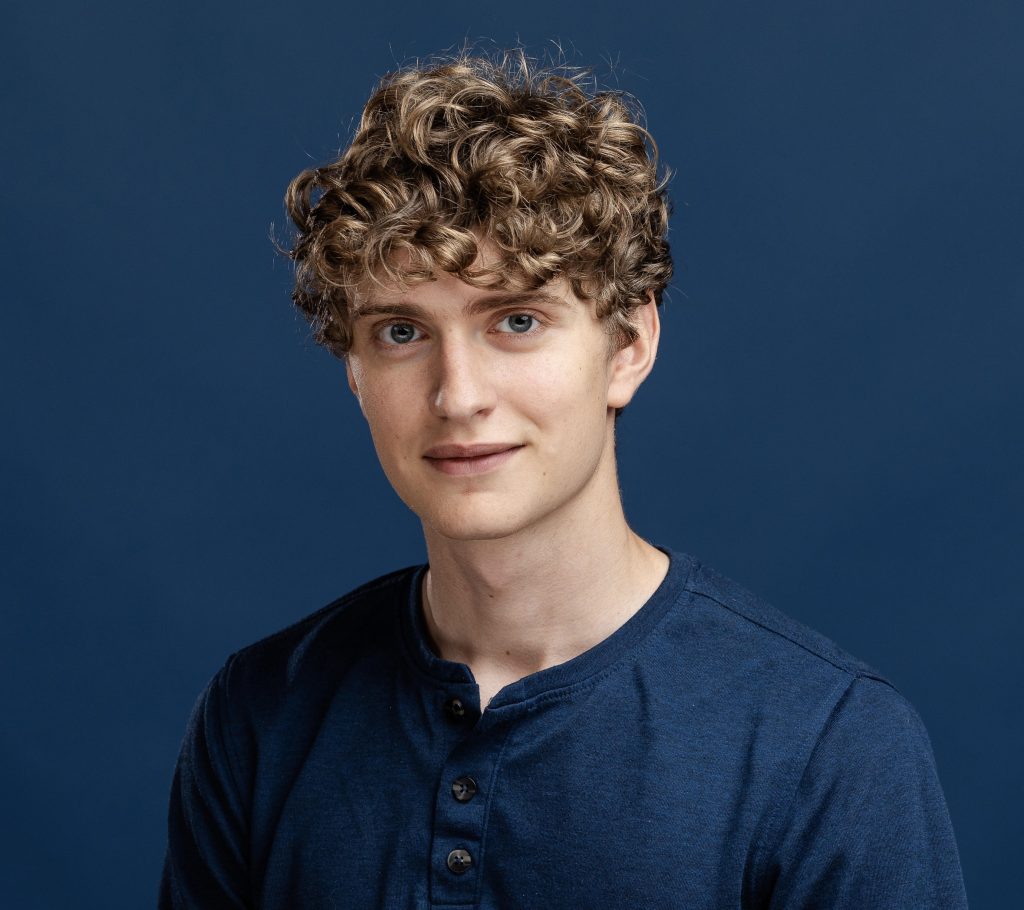Headshot of actor Luke DeMena, a young man with blond curly hair and blue eyes, against a dark blue background. 