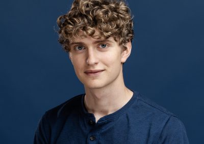 Headshot of actor Luke DeMena, a young man with blond curly hair and blue eyes, against a dark blue background.