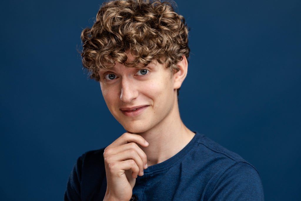 Headshot of Luke, a male actor with blond curly hair and blue eyes, smiling, against a dark blue background. 