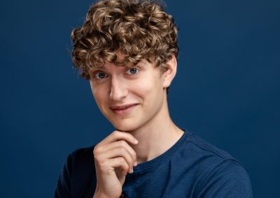 Headshot of Luke, a male actor with blond curly hair and blue eyes, smiling, against a dark blue background.
