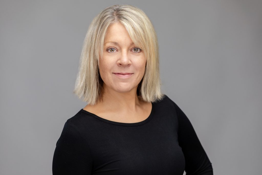 Headshot of a real estate agent. She is blond, wearing a black shirt, and smiling at the camera, against a grey backdrop.