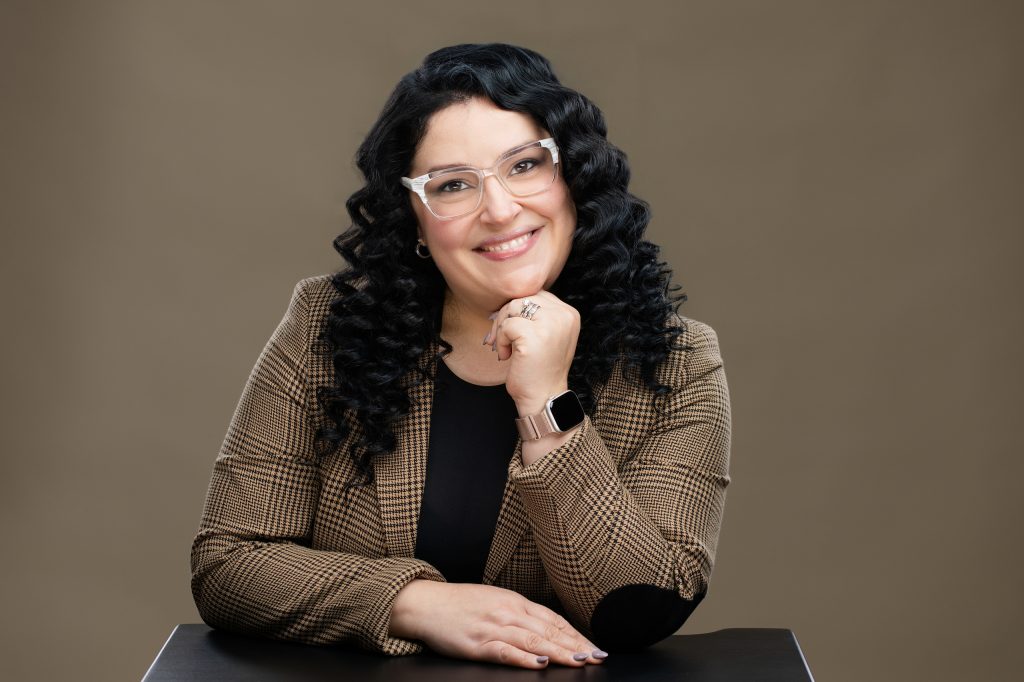 Professional woman's headshot, of a Latina woman resting her chin on her hand and smiling at the viewer.